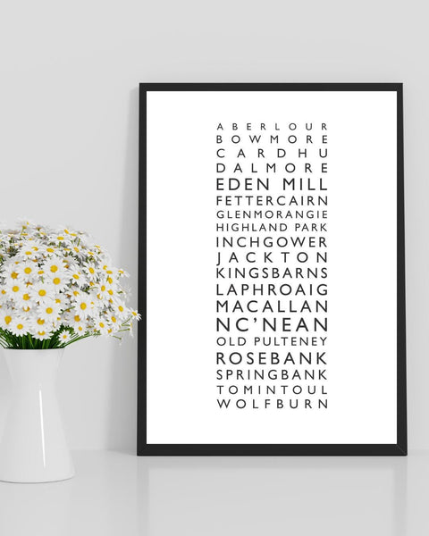 Minimalist framed wall print featuring a vertical list of Scottish whisky distilleries in clean black typography on a white background. The central text includes names like Aberlour, Bowmore, Cardhu, Dalmore, Eden Mill, Glenmorangie, Laphroaig, Macallan, and more. Styled with a neutral, Scandinavian-inspired aesthetic, the frame is surrounded by simple decor: a white vase with small blossoms on the left.
