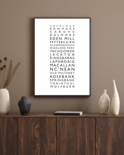 Minimalist framed wall print featuring a vertical list of Scottish whisky distilleries in clean black typography on a neutral background. The central text includes names like Aberlour, Bowmore, Cardhu, Dalmore, Eden Mill, Glenmorangie, Laphroaig, Macallan, and more. Styled with a neutral, Scandinavian-inspired aesthetic, the frame is surrounded by simple decor: a white vase with small blossoms on the left.