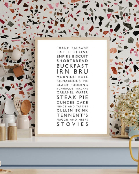 Minimalist framed wall print, displaying a vertical list of iconic Scottish foods and drinks in clean black typography on a white background. The design features items such as lorne sausage, tattie scone, Irn Bru, Buckfast, steak pie, Cullen skink, Tennent’s, and stovies. Styled in a neutral interior.