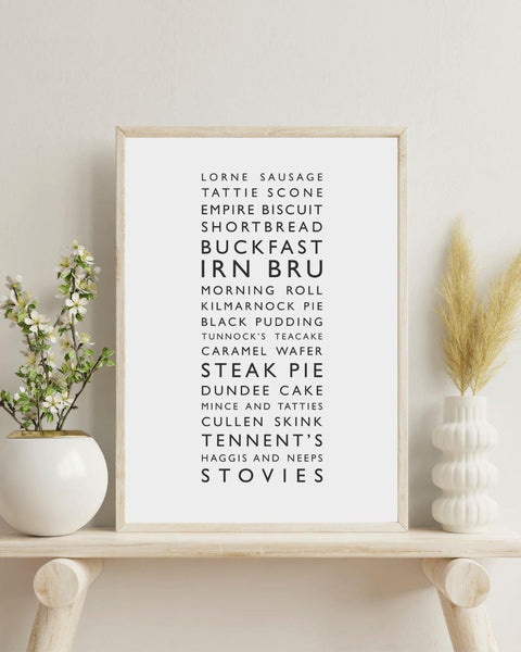 Minimalist framed wall print, displaying a vertical list of iconic Scottish foods and drinks in clean black typography on a white background. The design features items such as lorne sausage, tattie scone, Irn Bru, Buckfast, steak pie, Cullen skink, Tennent’s, and stovies. Styled in a neutral interior.