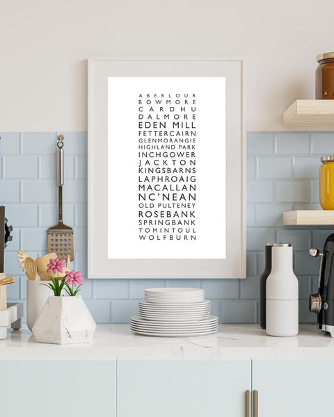 Minimalist framed wall print featuring a vertical list of Scottish whisky distilleries in clean black typography on a white background. The central text includes names like Aberlour, Bowmore, Cardhu, Dalmore, Eden Mill, Glenmorangie, Laphroaig, Macallan, and more. Styled with a neutral, Scandinavian-inspired aesthetic, the frame is surrounded by simple kitchen decor.